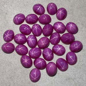 May include: A collection of oval-shaped, deep magenta star rubies. Each polished gemstone displays a six-rayed star pattern, with white lines radiating from the center. The rubies are scattered on a light grey surface.