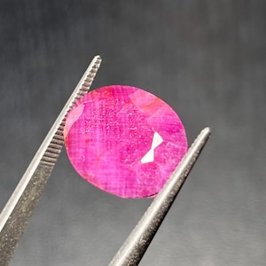 May include: A faceted, oval-shaped ruby gemstone held by metal tweezers. The ruby is a vibrant pinkish-red color, with visible internal textures and a reflective surface. The background is a dark gray.