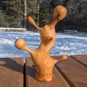 Peut inclure: Sculpture en bois faite à la main, avec une finition brun clair. La pièce d'art abstraite a une forme unique avec des protubérances arrondies s'étendant d'une forme centrale. La sculpture est exposée sur une surface en bois, avec un fond enneigé et des arbres.