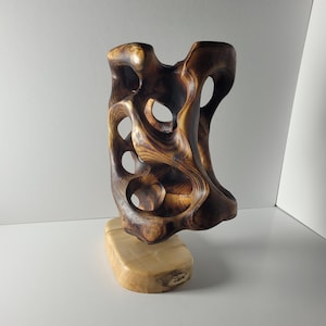 May include: A handcrafted wooden sculpture with a dark brown, organic form featuring multiple openings. The sculpture is mounted on a light-coloured, rectangular wooden base. The wood grain is visible, adding texture and detail to the artwork.