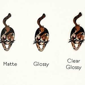 Puede incluir: Tres versiones de una pegatina con un gato calic&oacute; de dibujos animados con pelaje marr&oacute;n, negro y blanco. El gato est&aacute; en una pose juguetona, con la espalda arqueada y la cola levantada. Las pegatinas se muestran sobre un fondo blanco, con las palabras "Mate", "Brillante" y "Brillante transparente" impresas debajo de cada pegatina.