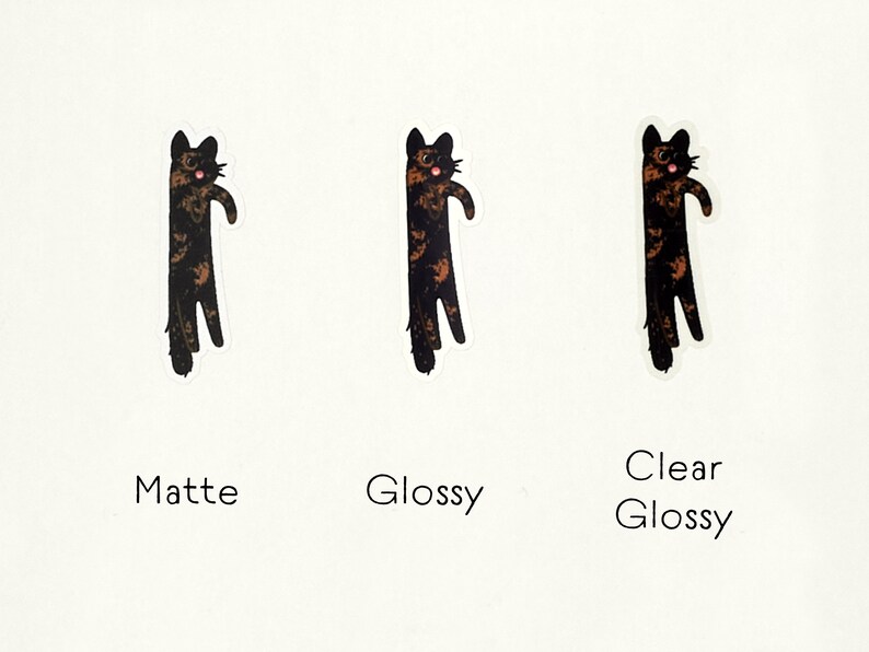 Op de afbeelding: Drie katten stickers, een met een matte afwerking, een met een glanzende afwerking en een met een heldere glanzende afwerking. Elke sticker toont een zwarte en oranje kat met een verraste uitdrukking.