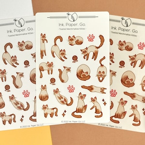 May include: Three sheets of stickers featuring illustrations of Siamese cats in various poses. The stickers are white with brown and pink accents. The text on the stickers reads "Ink. Paper. Go. - Toasted Marshmallow Kitties - 2022 Ink. Paper. Go. LLC".