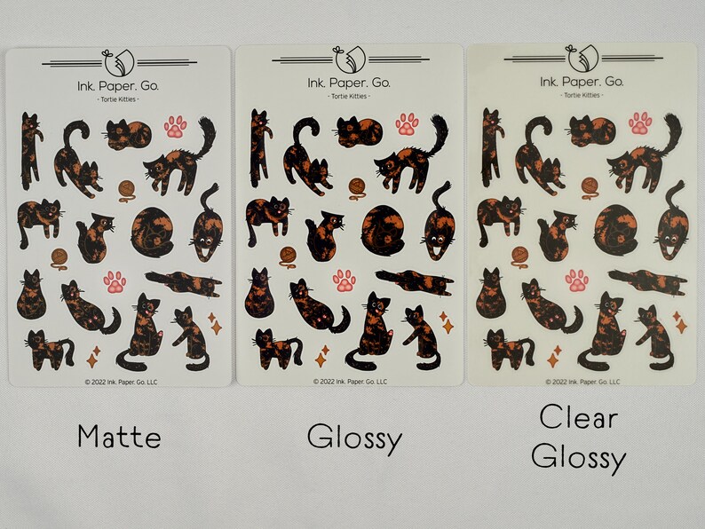 Op de afbeelding: Drie vellen met stickers met illustraties van schildpadkatten in verschillende poses. De stickers zijn gelabeld met "Mat", "Glans" en "Helder Glans". De tekst "Ink. Paper. Go. - Tortie Kitties" staat op elk vel.