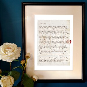 May include: A framed letter written in cursive script on aged paper. The letter is a personal correspondence with a handwritten seal in the bottom right corner. The letter is displayed against a dark blue wall with white flowers.