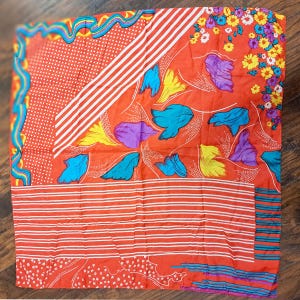 May include: A red silk scarf with a colorful geometric and floral pattern. The pattern includes stripes, polka dots, flowers, and abstract shapes in shades of blue, yellow, purple, and white.
