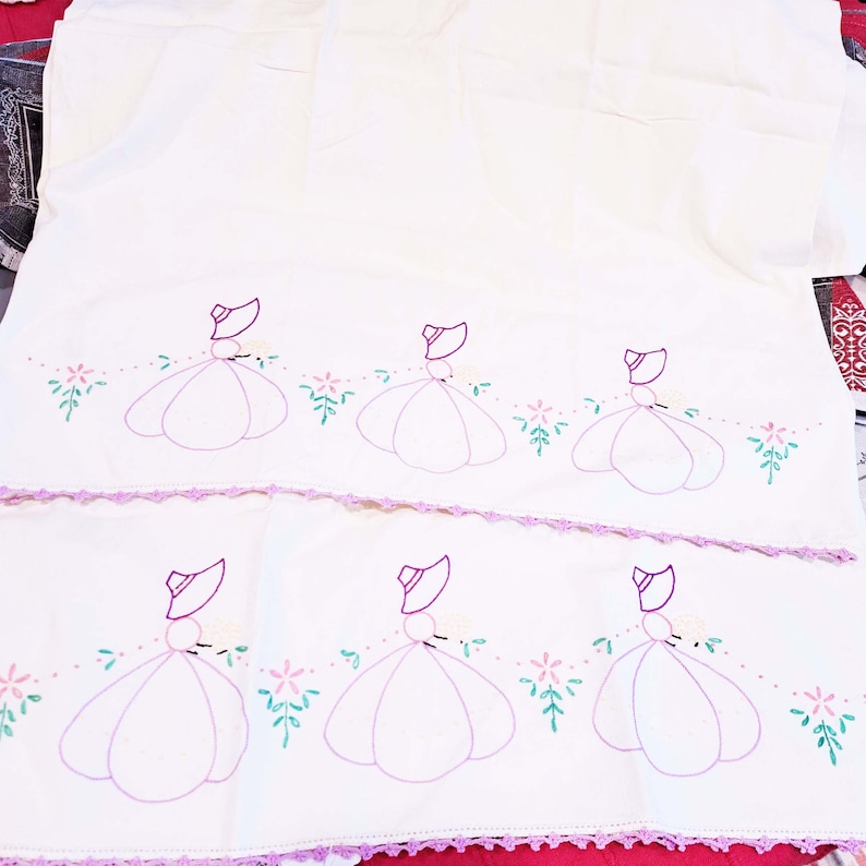 May include: Two white tea towels with embroidered purple and pink ladies in dresses and hats. The ladies are surrounded by green leaves and pink flowers. The towels have a crocheted edge.
