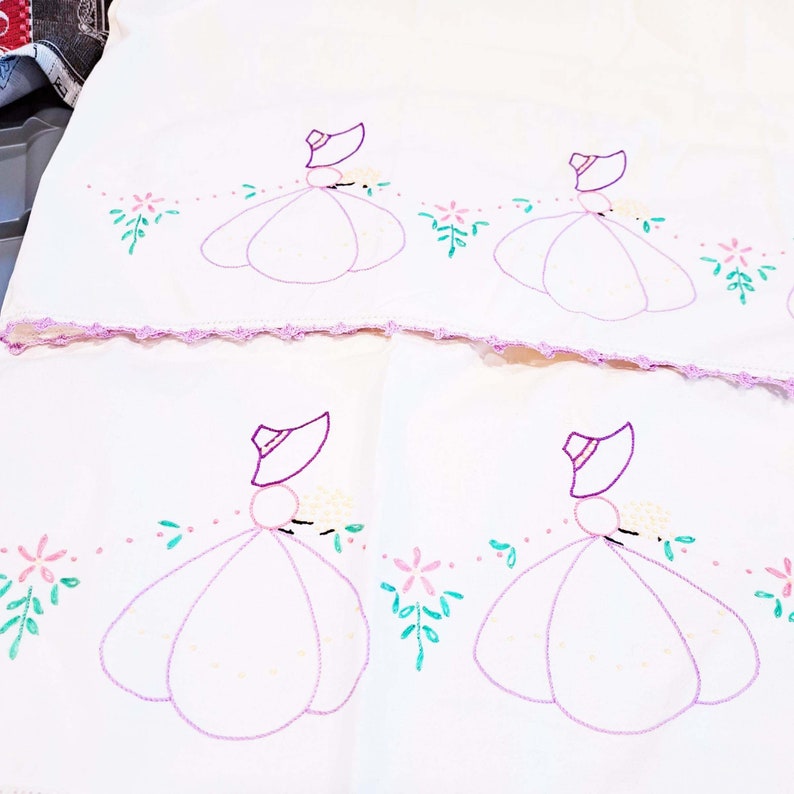 May include: Two white tea towels with embroidered purple ladies in pink dresses and green leaves. The ladies are wearing purple hats and holding white flowers. The towels have a crocheted edge.