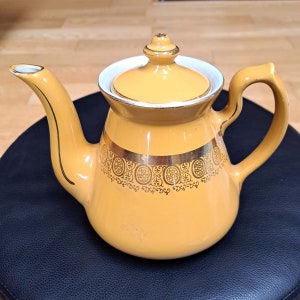 May include: A yellow teapot with a gold trim and a decorative pattern. The teapot has a lid and a handle.