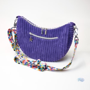 May include: A purple corduroy crescent-shaped handbag with a colorful, patterned strap. The bag has silver zippers and a star-shaped charm. The strap is a vibrant mix of colors, including red, blue, yellow, and green.