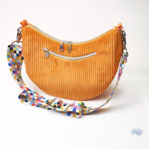 Puede incluir: Bolso de hombro de pana naranja con una correa estampada colorida. El bolso tiene forma curva, un compartimento principal con cremallera y un bolsillo frontal más pequeño con cremallera. Herrajes plateados y tirador de cremallera con carita sonriente.