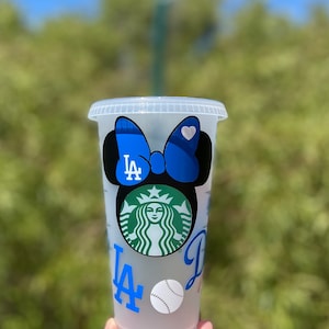 May include: A clear plastic reusable cup with a black Mickey Mouse ears design. The ears have a blue bow with a white heart on it. The cup has a Starbucks logo and the letters LA and a baseball.