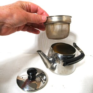 May include: A stainless steel tea kettle with a black handle and lid, and a removable mesh infuser. The lid has a black knob. The infuser is being held above the kettle.