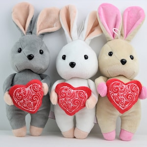 May include: Three soft toy rabbits in grey, white, and beige, each clutching a red heart with white embroidered patterns. The bunnies have pink or beige inner ears, set against a white backdrop.