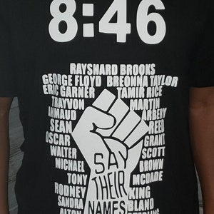 Può includere: T-shirt nera con scollo a V con la scritta "8:46" in bianco. Sotto, un pugno bianco e le parole "Say Their Names" sono visualizzate. I nomi delle persone sono elencati attorno al pugno.