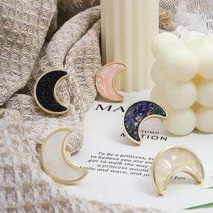 Puede incluir: Cuatro broches de media luna de oro con incrustaciones de diferentes colores. Las incrustaciones son negras con purpurina, rosas, azules y blancas.