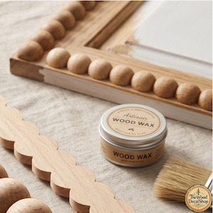 Op de afbeelding: Een ronde metalen blik met Artisan houtwas met een beige etiket, een houten frame met een kralenrand, een apart houten sierlijststuk met een geschulpte rand en een kwast met natuurlijke haren. Het etiket bevat de tekst Artisan WOOD WAX.