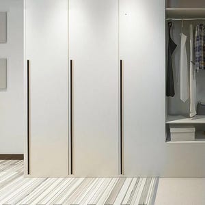 Könnte beinhalten: Ein weißer Kleiderschrank mit drei hohen Türen und langen, vertikalen schwarzen Griffen. Die rechte Seite des Schranks ist offen und zeigt eine Kleiderstange mit verschiedenen Kleidungsstücken, ein Regal und eine Aufbewahrungsbox. Der Boden ist hell gestreift.