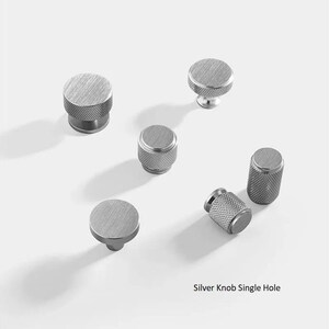 May include: Six silver cabinet knobs with a single hole for installation. The knobs have a textured, brushed finish and are round or cylindrical in shape.
