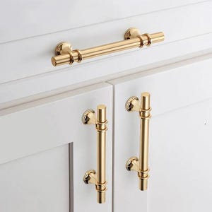 May include: Gold cabinet hardware set featuring a pull handle and two knobs. The pull handle is long and straight with a cylindrical design. The knobs are round with a cylindrical design.