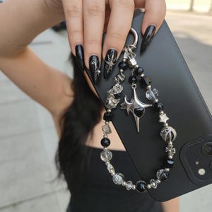 May include: Black phone case with a beaded charm. The charm features black and clear beads, silver-toned metal accents, and a star pendant with a heart. The phone case is held by a person with long black nails.
