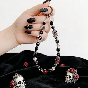 May include: A beaded keychain with black, red, and silver beads, including skull and rose charms. The keychain is held against a black velvet background, with additional skull and rose decorations.