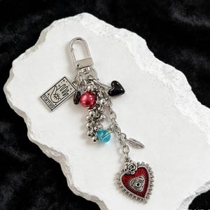 May include: A silver keychain with a variety of charms. Charms include a red heart with an eye design, a small card with the word "FORTUNE", a black heart, and various beads. The keychain is on a white, textured surface.