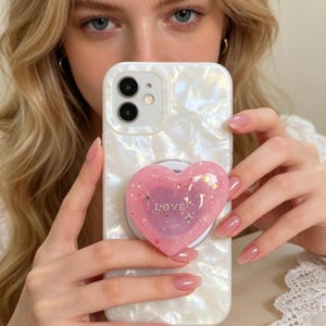 May include: White pearlescent phone case with a pink heart-shaped phone grip. The grip has the word "LOVE" and glitter accents. The phone case has a dual-lens camera and flash.