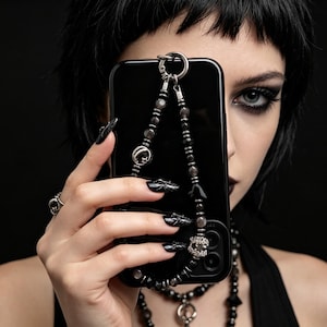 May include: Black smartphone case with a beaded strap. The strap features dark gray beads, a silver-tone charm, and a decorative gem. The case is held in front of a person's face, with black nail polish and jewelry visible.