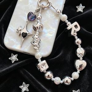 May include: A phone charm featuring a mix of silver-toned metallic beads, white pearls, and a letter R charm. It includes a dangling star-shaped pendant with a black heart center and a small purple heart charm, all attached to a white phone case.
