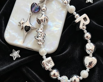 May include: A phone charm featuring a mix of silver-toned metallic beads, white pearls, and a letter R charm. It includes a dangling star-shaped pendant with a black heart center and a small purple heart charm, all attached to a white phone case.
