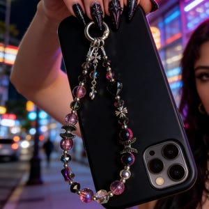 May include: Black phone case with a beaded charm strap. The strap features a mix of pink, purple, and clear beads, along with silver accents and charms. The phone case has a silver clasp for attaching the strap.