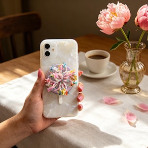 May include: A white pearlescent smartphone case featuring a circular lavender phone grip adorned with a cluster of colorful, multi-shaped beads and small flower charms in shades of pink, yellow, blue, and green attached to the center back.