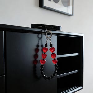 May include: A black phone case with a silver keyring attached to a beaded lanyard. The lanyard features red heart-shaped beads and black round beads. The phone case is attached to a black cabinet.