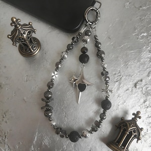 May include: A beaded phone charm with a silver star pendant featuring a heart, and various dark gray beads. The charm is attached to a silver clasp. Two decorative metal objects are also visible.