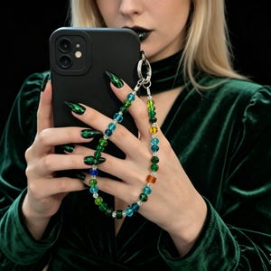 May include: Black phone case with a colorful beaded phone strap. The strap features a variety of colored beads, including green, blue, yellow, and orange. The phone case is attached to a silver-colored clasp.