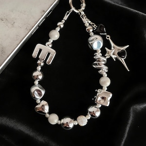 May include: A silver-toned keychain with a variety of beads and charms. The keychain features an "E" charm, round and irregular silver beads, white pearls, and star and heart charms. The background is a black, silky fabric.