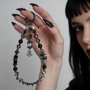 Könnte beinhalten: Ein schwarzer Schlüsselanhänger mit Perlen und verschiedenen Anhängern, darunter eine Fleur-de-Lis, geometrische Formen und florale Designs. Der Schlüsselanhänger hat einen großen schwarzen Ring und ist mit schwarzen, klaren und lila Perlen verziert. Die Gesamtästhetik ist gotisch und kantig.