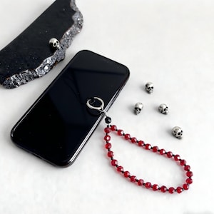 Puede incluir: Un teléfono inteligente negro con una correa de muñeca de cuentas rojas. La correa presenta pequeñas cuentas rojas facetadas y cuentas negras. Alrededor del teléfono hay pequeños dijes de calaveras plateadas.