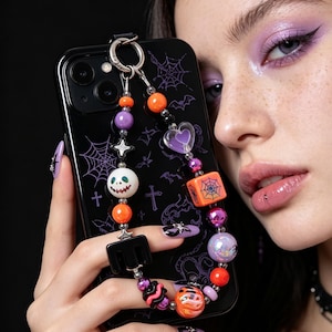 May include: A black smartphone case featuring a purple spiderweb and bat pattern, paired with a decorative beaded phone charm. The charm includes orange, purple, and iridescent beads, a heart-shaped bead, a square spiderweb bead, and a skull bead.