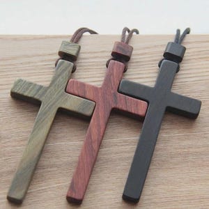 May include: Three wooden cross pendants on dark brown cords. The crosses are made of wood in three distinct colors: a light olive green with visible grain, a reddish-brown with dark streaks, and a solid matte black finish with a smooth texture.