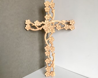 May include: Wooden cross with a floral design. The cross is made of light-colored wood and features carved flowers and vines. The flowers are arranged along the arms and stem of the cross, creating a decorative and elegant look.
