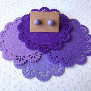 May include: A pair of purple stud earrings on a brown card, displayed on a purple doily.