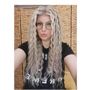 Könnte beinhalten: Ein schwarzes T-Shirt mit weißem Text auf der Brust, kombiniert mit herzförmigen Brillen mit dünnen schwarzen Rahmen. Der Look wird durch langes, welliges, platinblondes Haar mit dunkleren Ansätzen ergänzt, das in einem Mittelscheitel fällt.