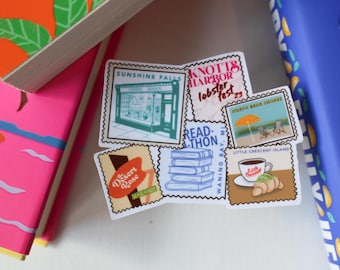 May include: A collection of travel-themed stickers with various designs, including a bookstore, a lobster festival, a beach scene, and a coffee cup with a croissant. The stickers are arranged near colorful books.