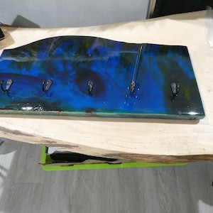 May include: A rectangular wall-mounted key holder with a wavy top edge. The holder features a vibrant blue and green resin design with four metal hooks. The resin art resembles an abstract ocean scene, adding a decorative touch to any entryway.