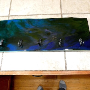 May include: A rectangular wall-mounted key holder with five black metal hooks. The wooden base is painted with a swirling pattern of blue, green, and black. The key holder is designed for home organization.