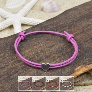Puede incluir: Un brazalete de cordón de cuero rosa con un dije de corazón plateado. El brazalete está atado con un nudo en un extremo y tiene un dije de corazón plateado en el otro extremo.
