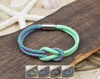 Könnte beinhalten: Ein Armband mit einem zweisträngigen Kordeldesign und einem zentralen Knoten. Eine Seite der Kordel ist hellgrün, die andere Seite ist dunkelblaugrün mit kleinen rosa und hellblauen Sprenkeln. Es hat einen zylindrischen, silberfarbenen Metallverschluss.