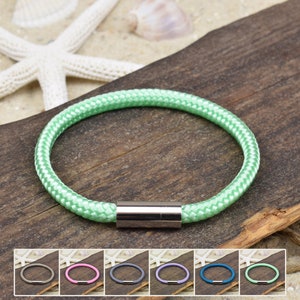 Könnte beinhalten: Ein grünes Seil-Armband mit einem silbernen Magnetverschluss. Das Armband liegt auf einer Holzoberfläche mit einem Seestern und Muscheln.